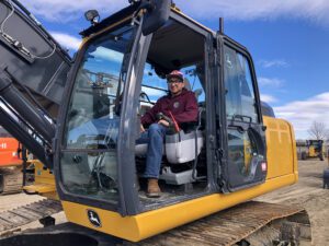 Operator sits in the cab of a yellow construction excavator at a job site, wearing a maroon hoodie and striped cap.