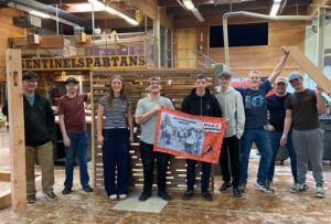 Nine students stand in a woodshop posing for a group photo, holding an orange banner in front of wooden frames and tools.