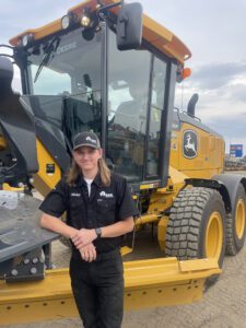 Young man in a black RDO uniform standing beside a yellow John Deere construction machine on a dirt lot, smiling at the camera.