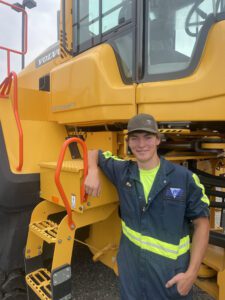 Wade Heinzman - Build Montana Smiling worker in high-vis blue coveralls leans on a yellow Volvo heavy machine nearby a red handrail.